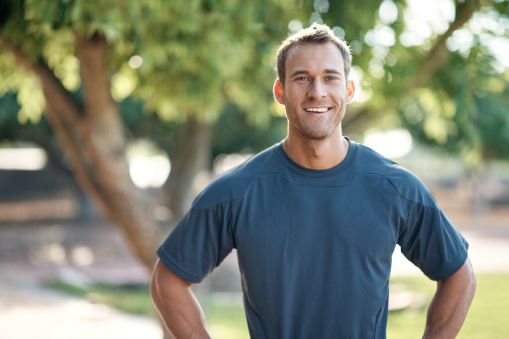 A man standing confidently in a park with his arms at his sides.