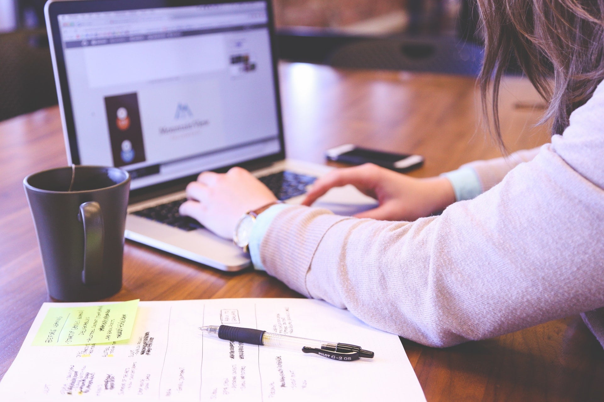 A woman working on a laptop with a coffee cup near by as well as a pen and paper