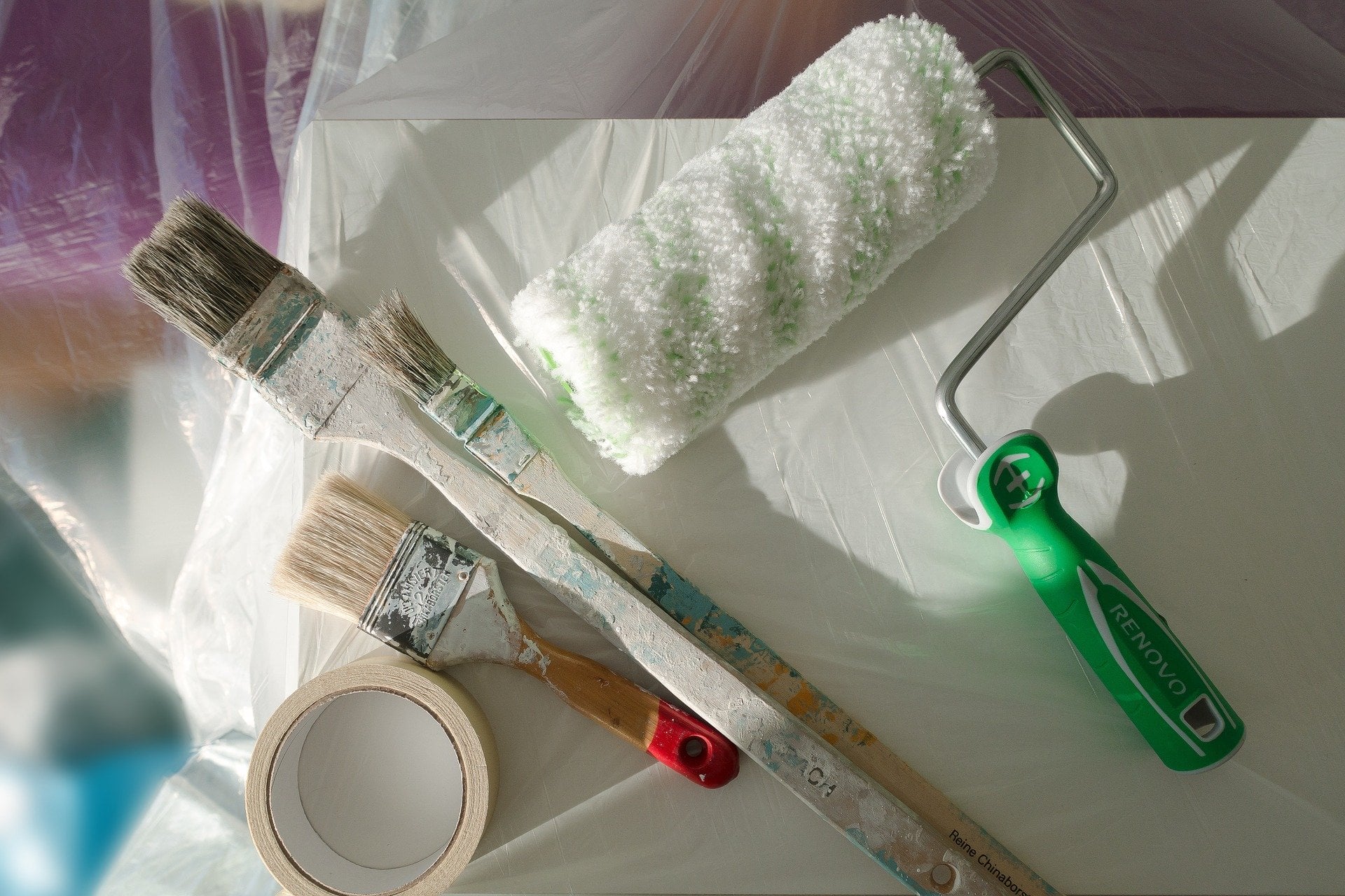 A paint roller sits alongside three paint brushes and some painters tape atop a table covered in a drop cloth.
