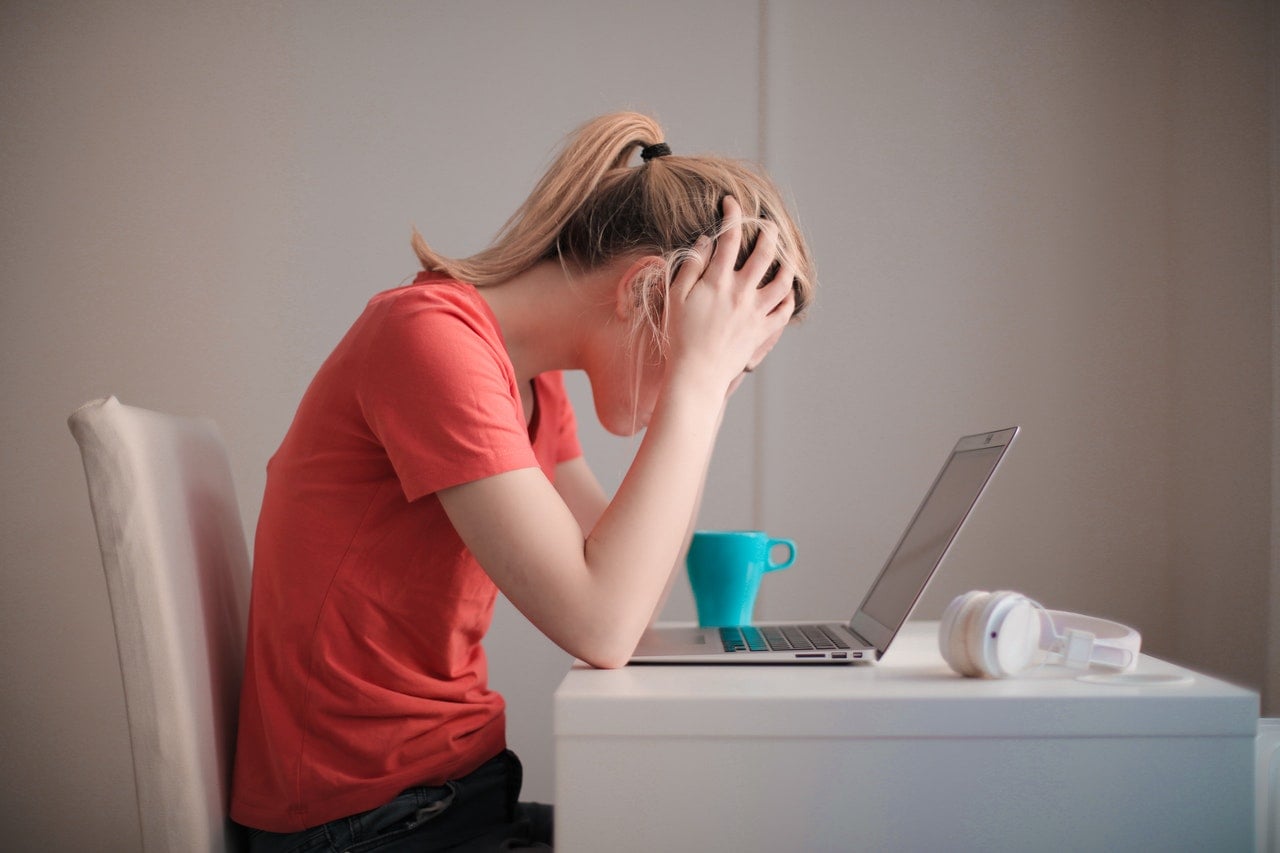 A stressed out woman sits at a desk with her hands on her head while looking at an open laptop.