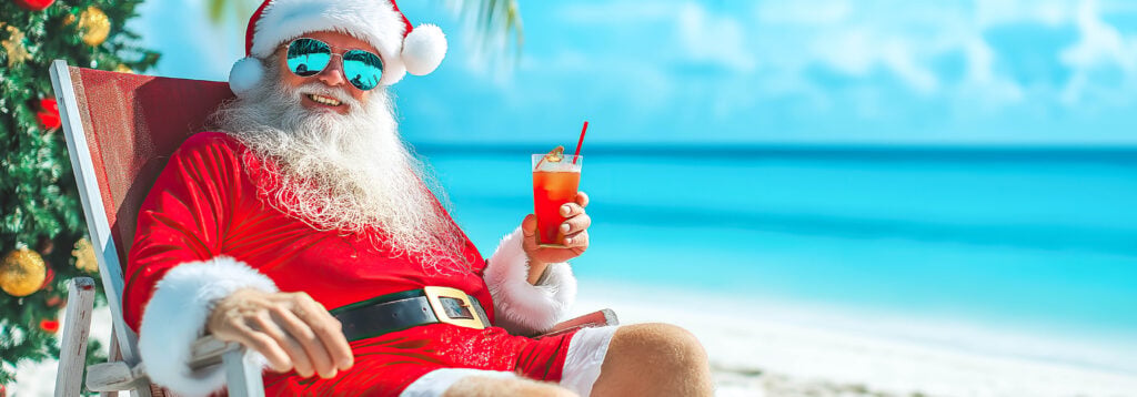 A smiling Santa Claus is relaxing on a tropical beach with a drink in his hand.