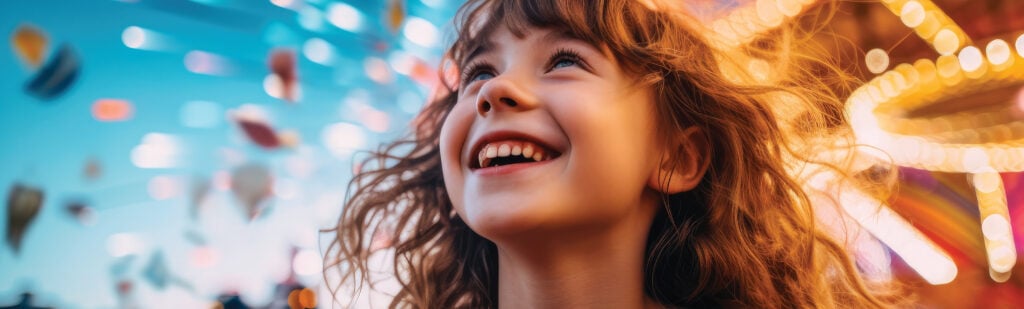 A smiling child is looking up at the lights at a fairground.